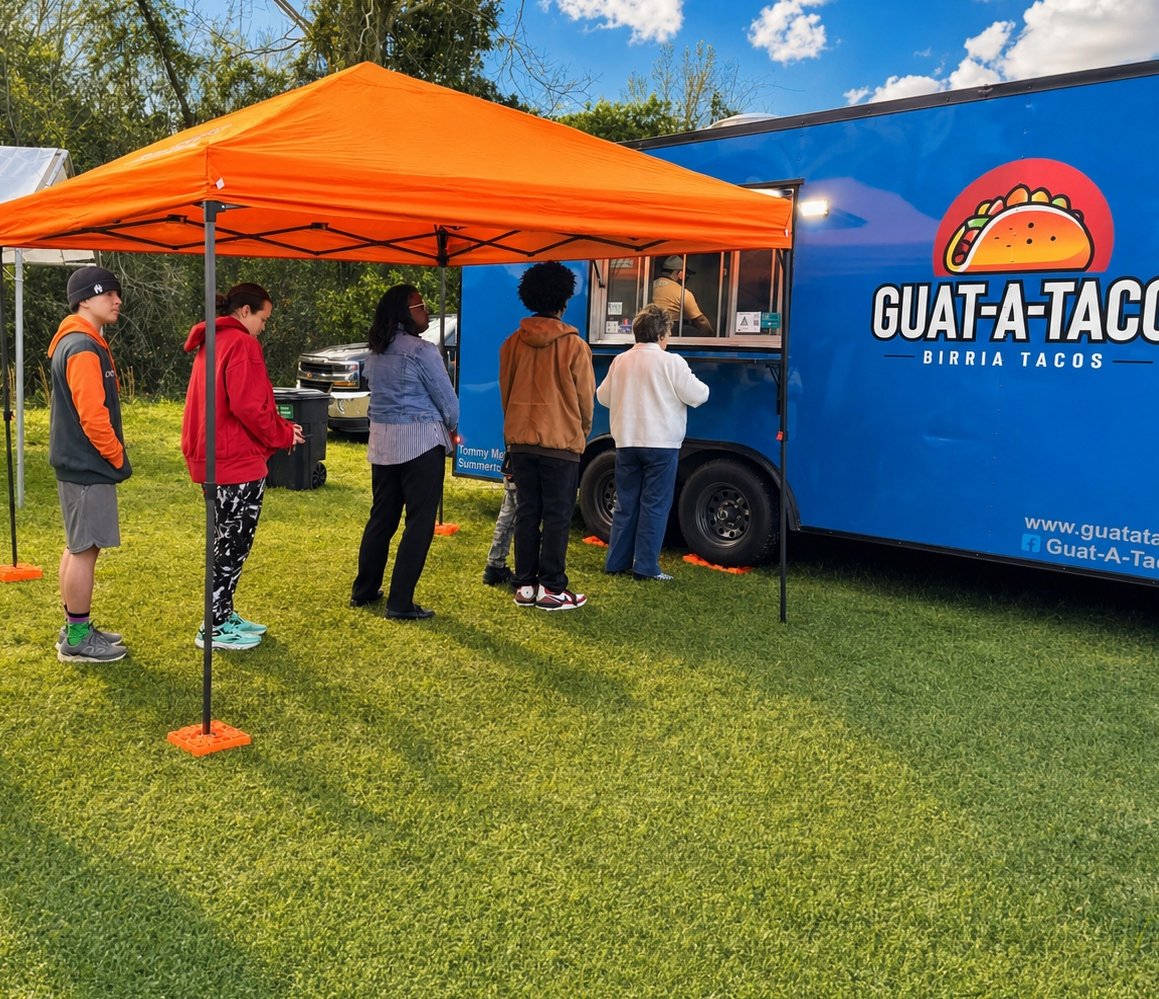 A line of guests waiting under an orange canopy in front of the blue Guat-A-Taco food truck in Manning, SC — sunny afternoon, green field, birria tacos logo on the truck